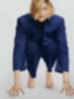 Greta Gerwig, with shoulder-length hair and painted fingernails and toenails, and wearing a suit, crouches and looks up